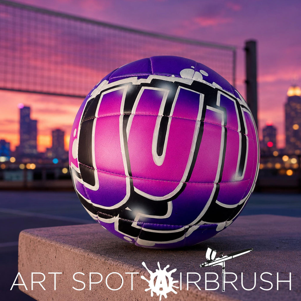 A purple and black custom volleyball personalized with the word "JUJU" painted on it, sitting on a concrete surface against a city skyline at sunset.