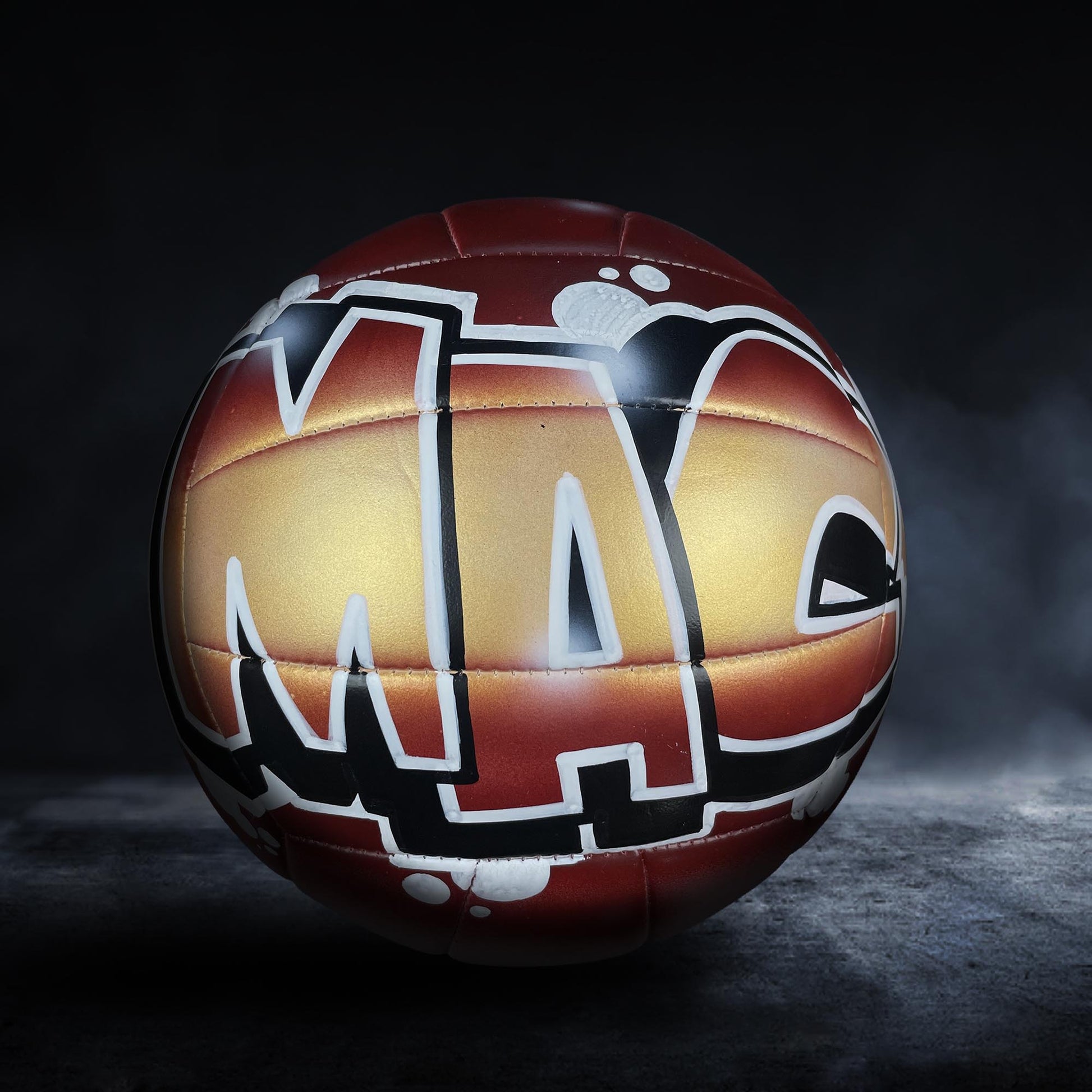 A red and gold soccer ball with the word "MA" painted on it, set against a dark background with a misty effect.