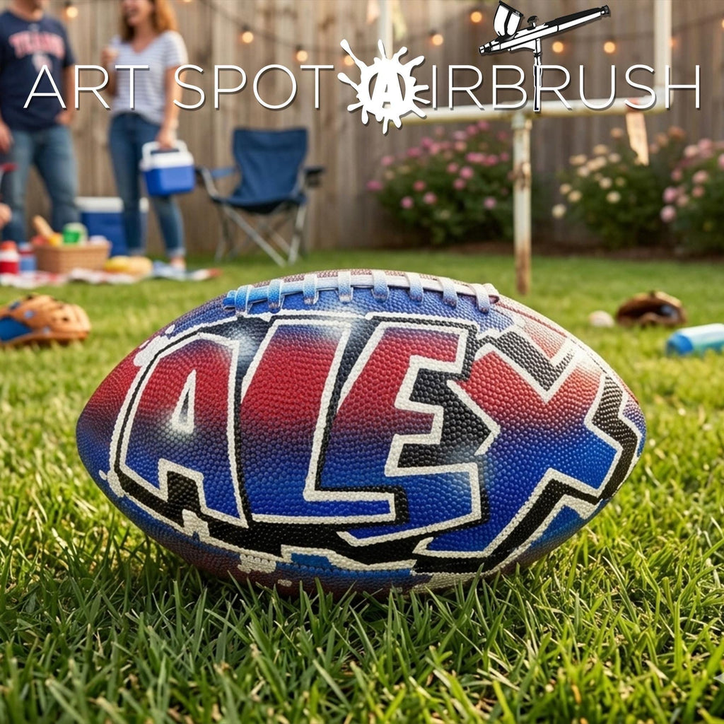 A custom football personalized with the name "ALEX" painted on it, sitting on a grassy field with a wooden fence and some people in the background.