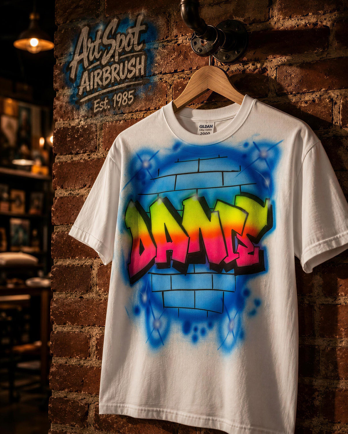 A white t-shirt with a colorful graffiti design on it, hanging on a wooden hanger against a brick wall.