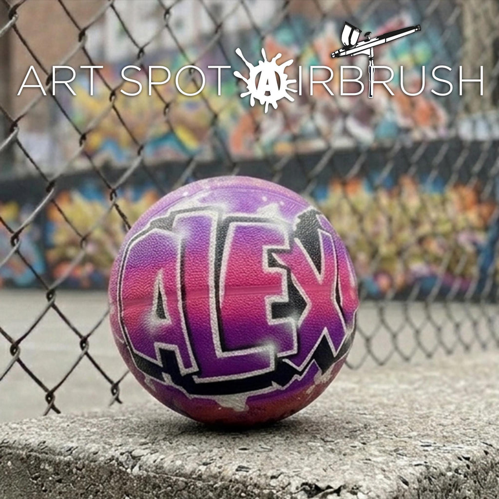 A purple basketball with the word "ALEX" painted on it, sitting on a concrete surface in front of a chain-link fence with graffiti on it.