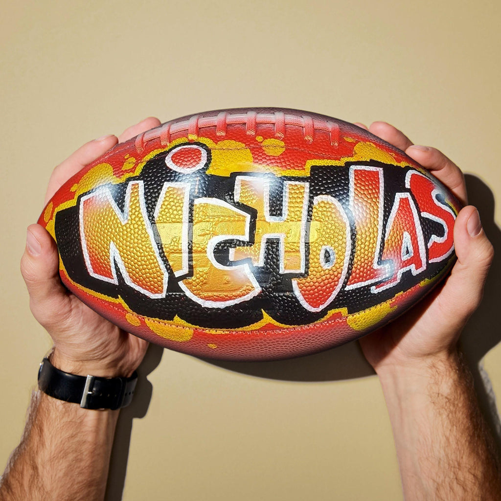 A person's hands holding a custom football personalized with the name "NICHOLAS" painted on it in graffiti-style lettering.