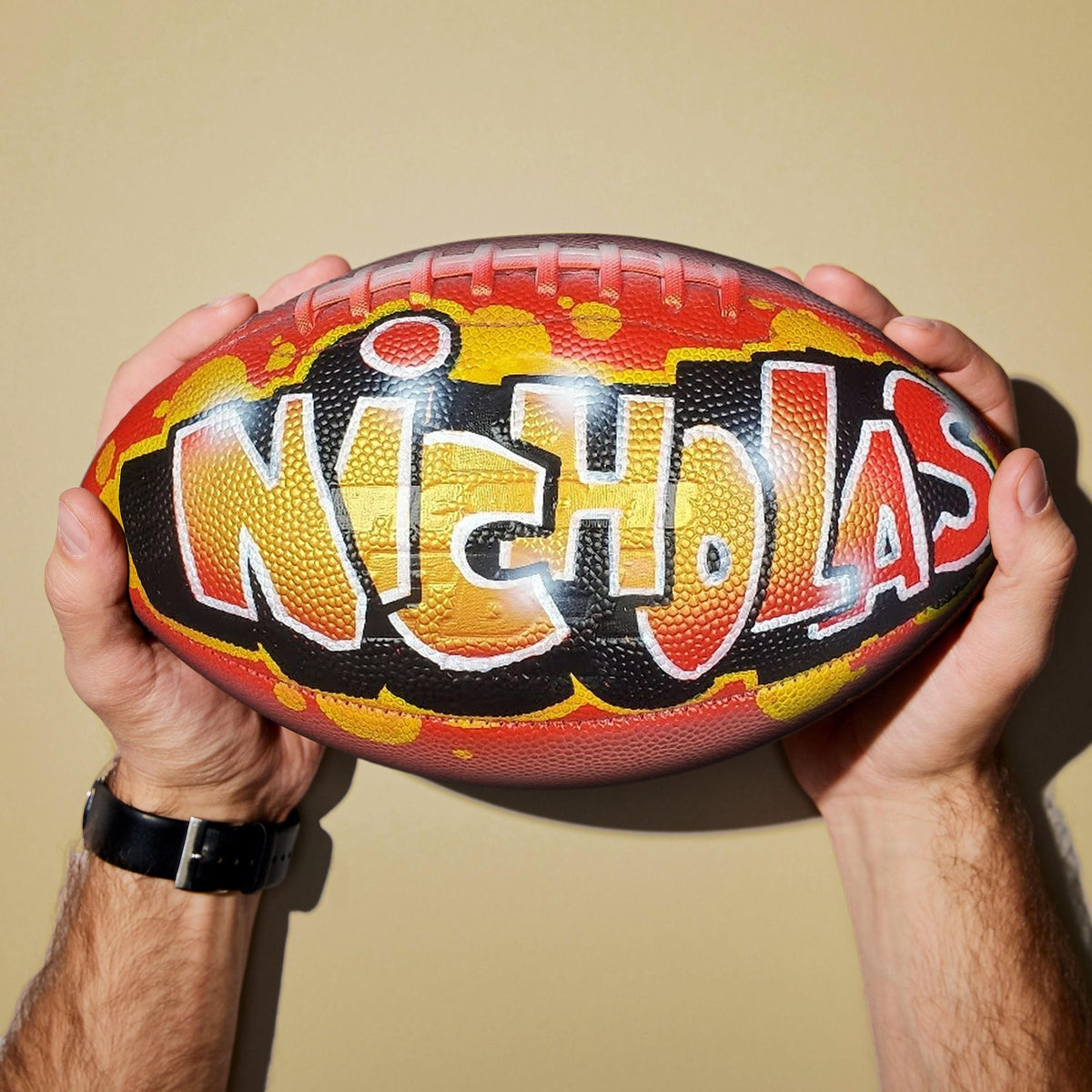 A person's hands holding a custom football personalized with the name "NICHOLAS" painted on it in graffiti-style lettering.