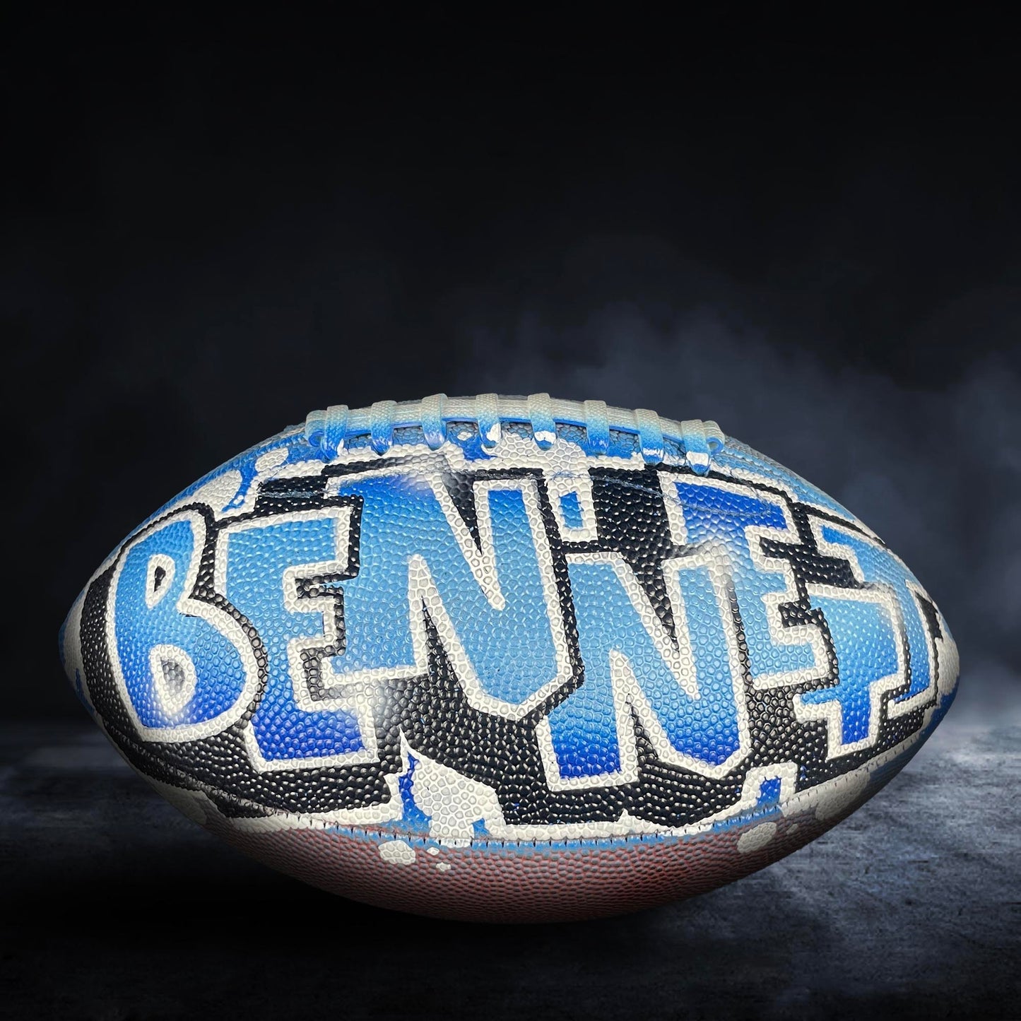 A football with the name "BENNET" painted on it, against a dark background with smoke or fog.