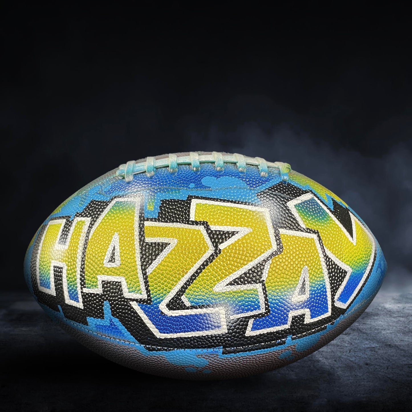 A colorful football with the word "HAZY" painted on it, sitting on a dark surface with smoke or fog in the background.