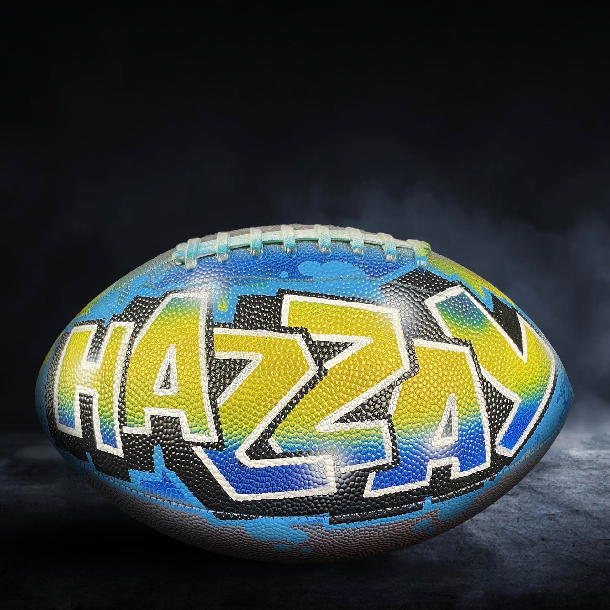 A colorful football with the word "HAZY" painted on it, sitting on a dark surface with smoke or fog in the background.