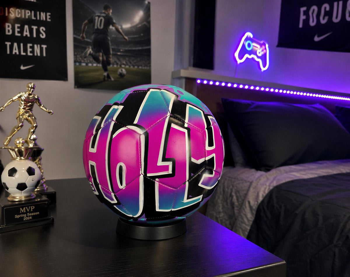 A brightly colored soccer ball with the word "HOLY" painted on it, placed on a table in a bedroom with posters and a neon sign in the background.