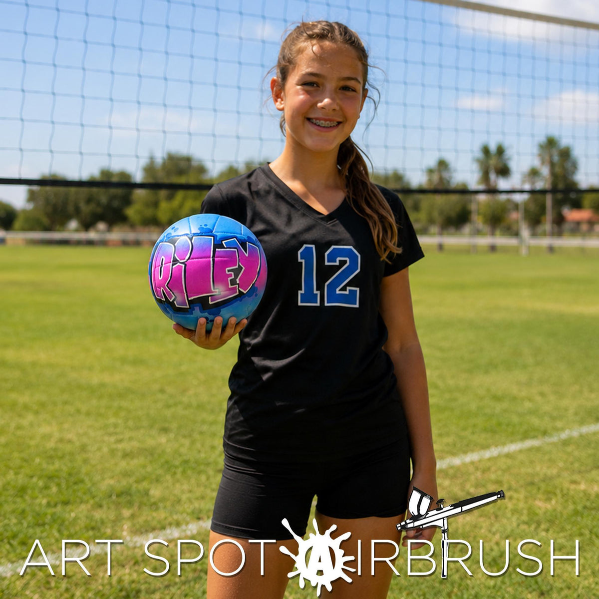 A young girl in a black jersey with the number 12 is holding a personalized custom volleyball with the word "Riley" on it, standing on a grassy field with a volleyball net in the background.