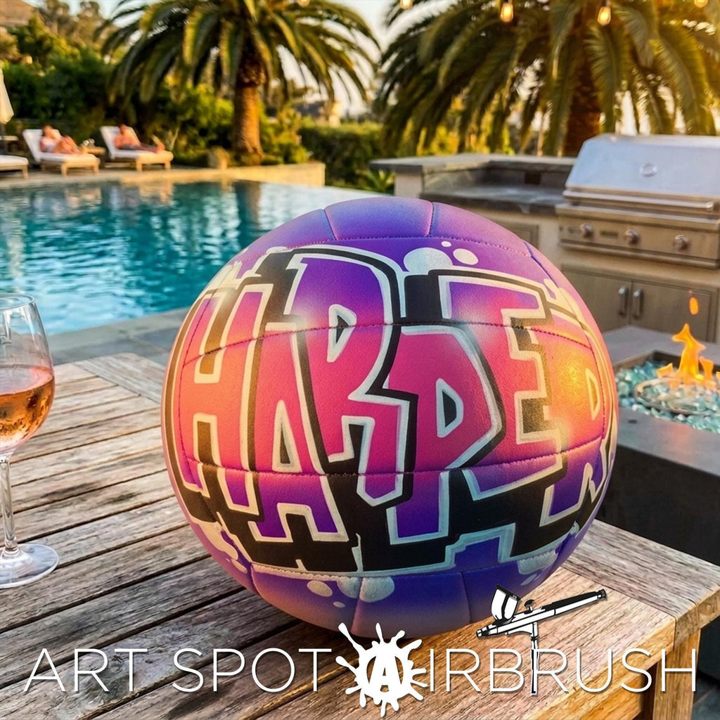 A purple and pink custom volleyball personalized with the word "HARPER" on it, placed on a wooden table near a swimming pool and a grill.