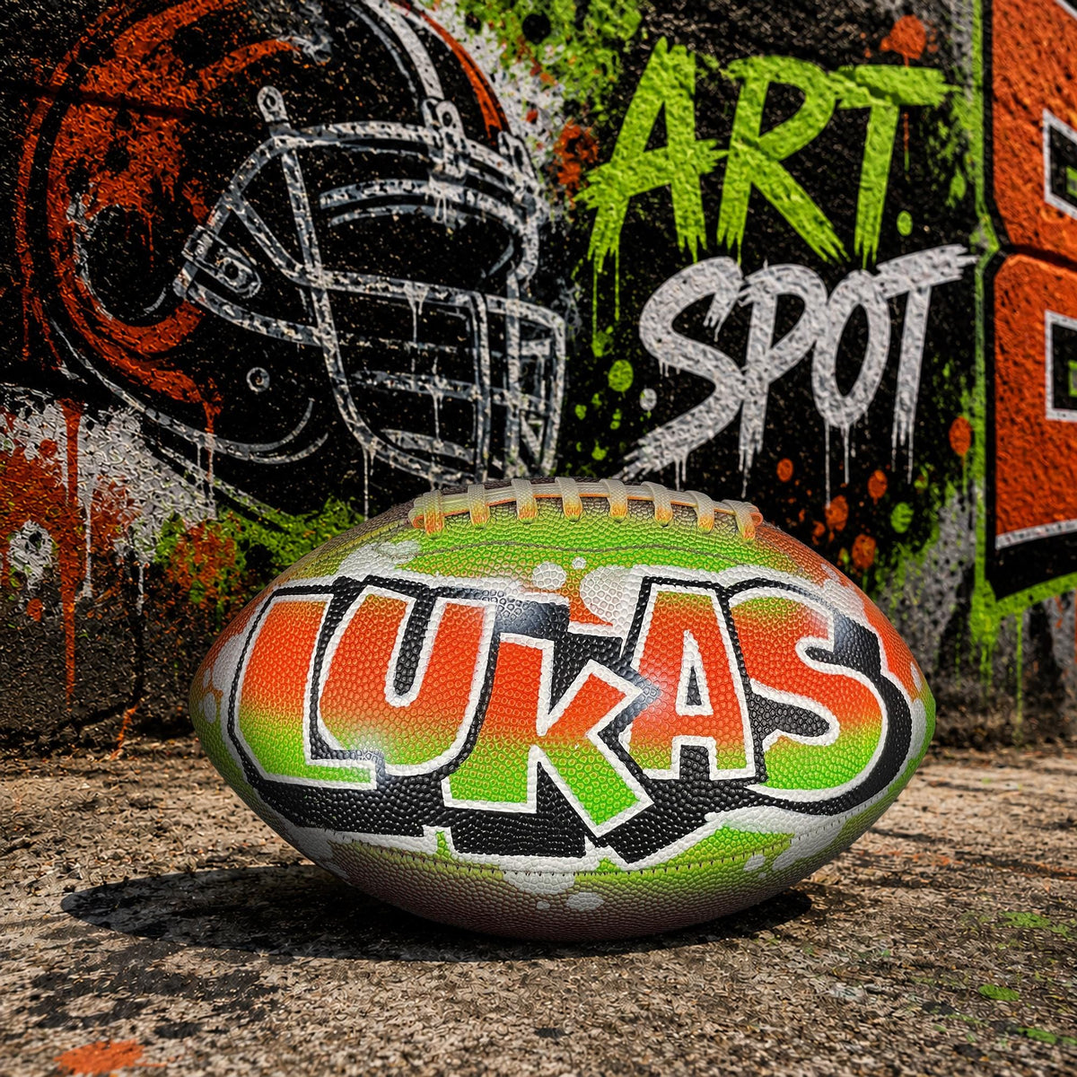 A personalized custom football with the name "LUKAS" painted on it, sitting on a concrete surface in front of a graffiti-covered wall.