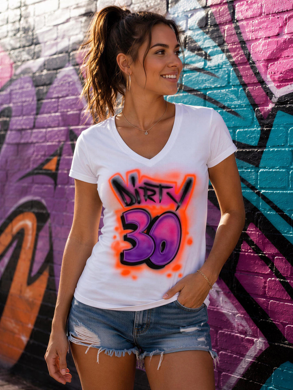 A young woman wearing a white t-shirt with a graffiti design and denim shorts stands in front of a colorful graffiti wall.