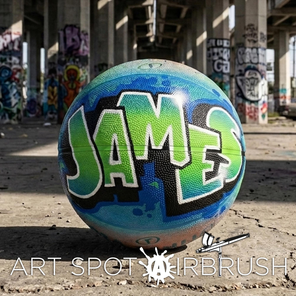 A colorful basketball with the name "JAMES" painted on it, sitting on a concrete surface with graffiti in the background.