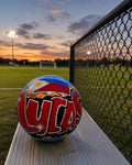 A soccer ball with the word "ULA" written on it, sitting on a bench in a park at sunset.