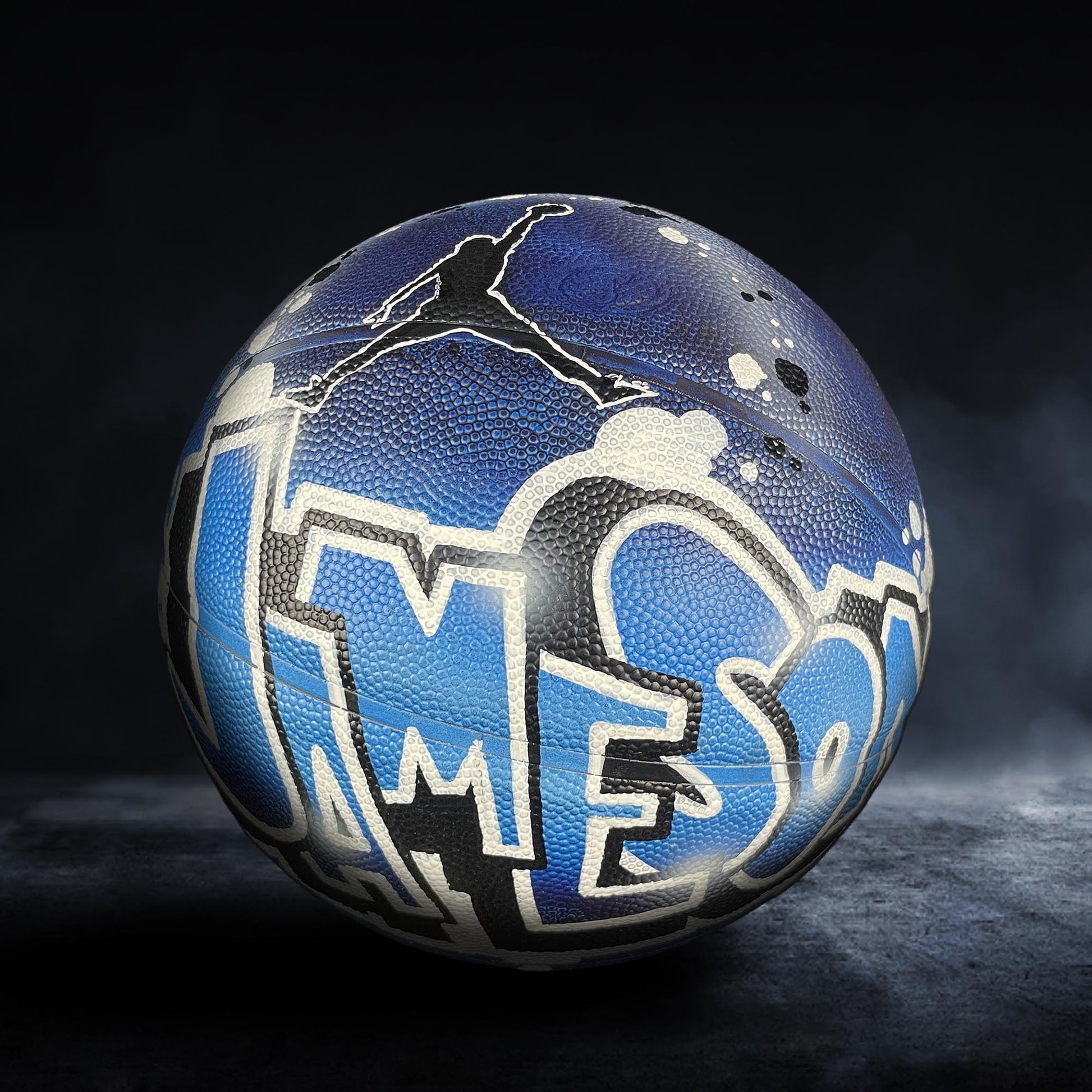 A basketball with a blue and white design featuring the word "JAMES" on it, sitting on a dark surface.
