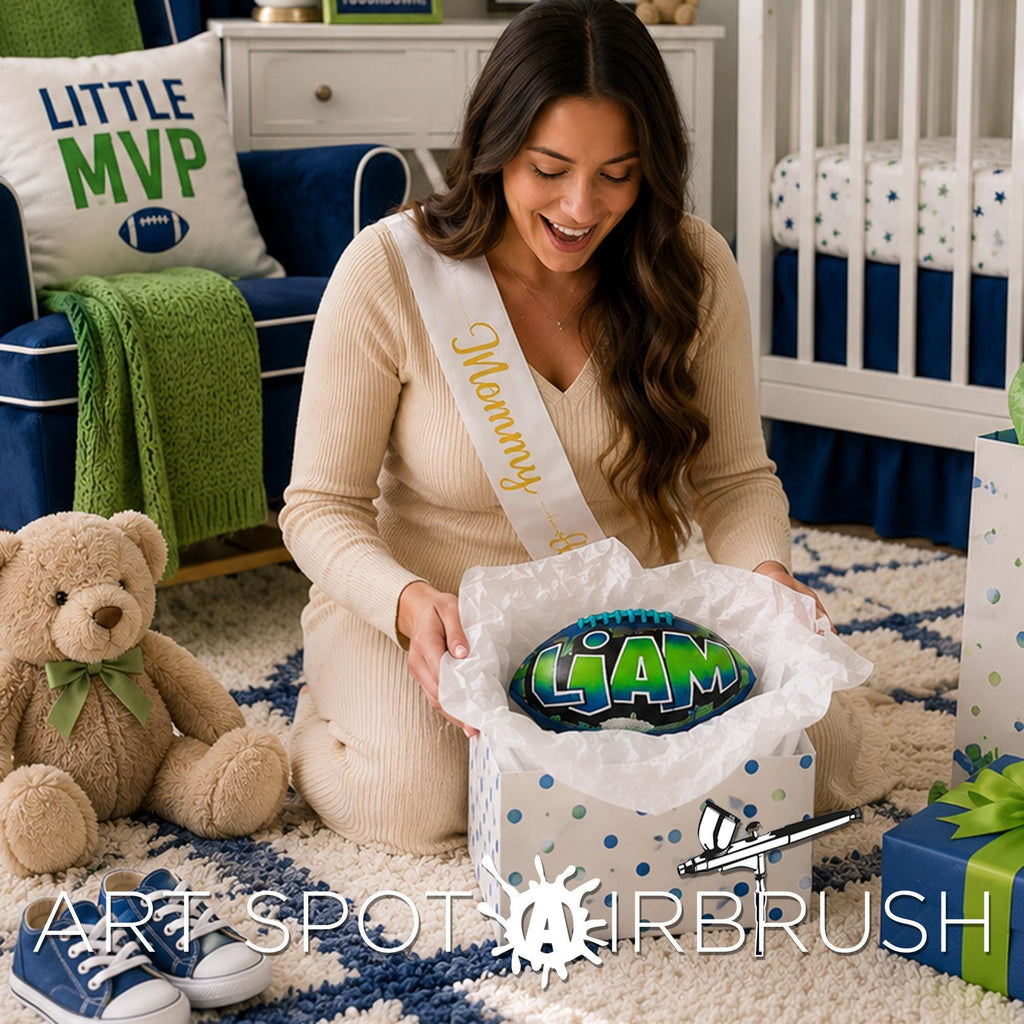 A woman in a beige sweater is kneeling on the floor, holding a gift box with a personalized custom football with the name LIAM. Teddy bear is sitting next to her, and there are other sports balls and a football-themed pillow in the room.