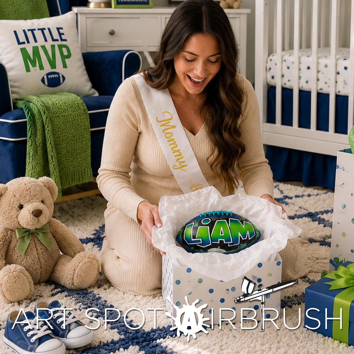 A woman in a beige sweater is kneeling on the floor, holding a gift box with a personalized custom football with the name LIAM. Teddy bear is sitting next to her, and there are other sports balls and a football-themed pillow in the room.