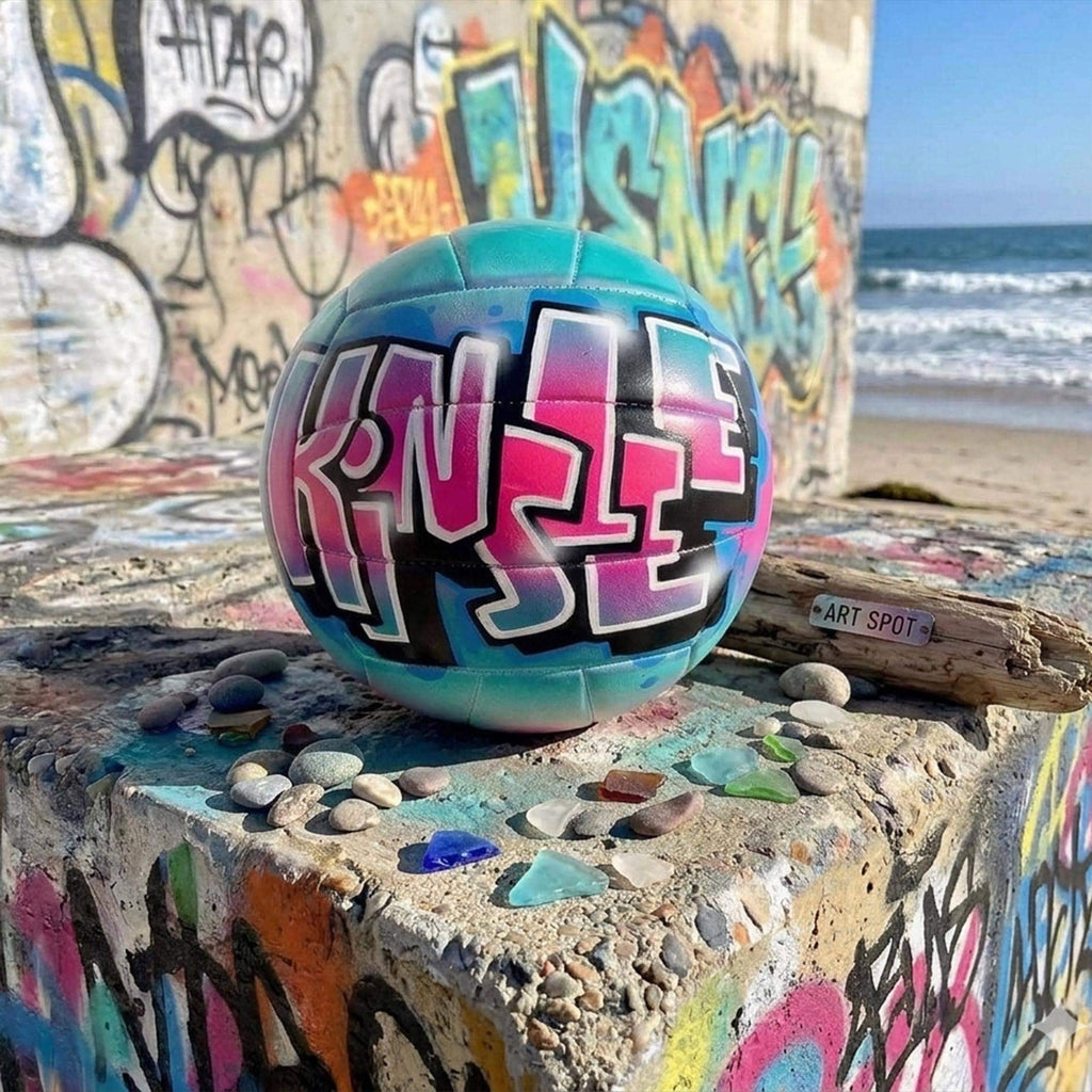 A colorful, graffiti-style custom volleyball personalized with the name "Kinsley" painted on it, placed on a concrete surface with various small rocks and pebbles scattered around it. In the background, there is a wall covered in vibrant graffiti.