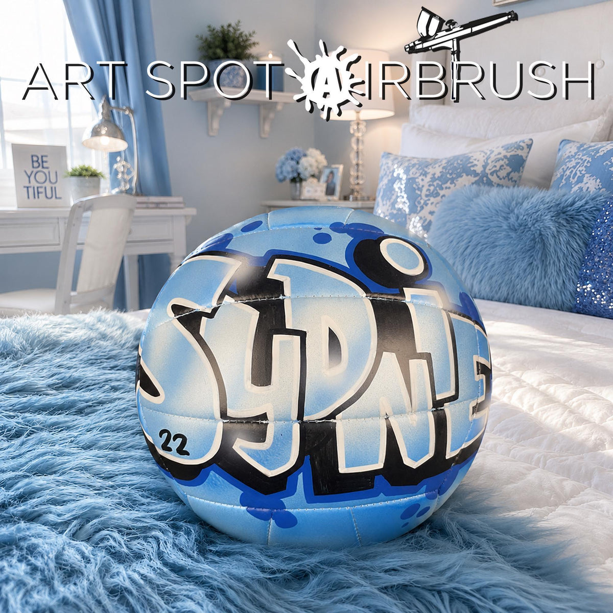 A blue and white custom volleyball with the word "Sydnie" painted on it, sitting on a bed with a fluffy blue blanket.