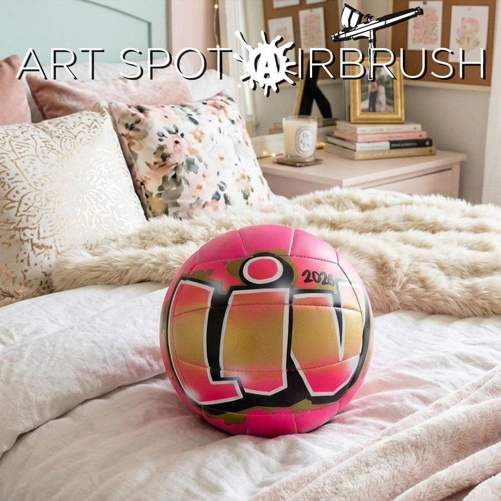 A pink and black A custom volleyball with the text LIV is placed on a bed with a floral pillow and a stuffed animal.