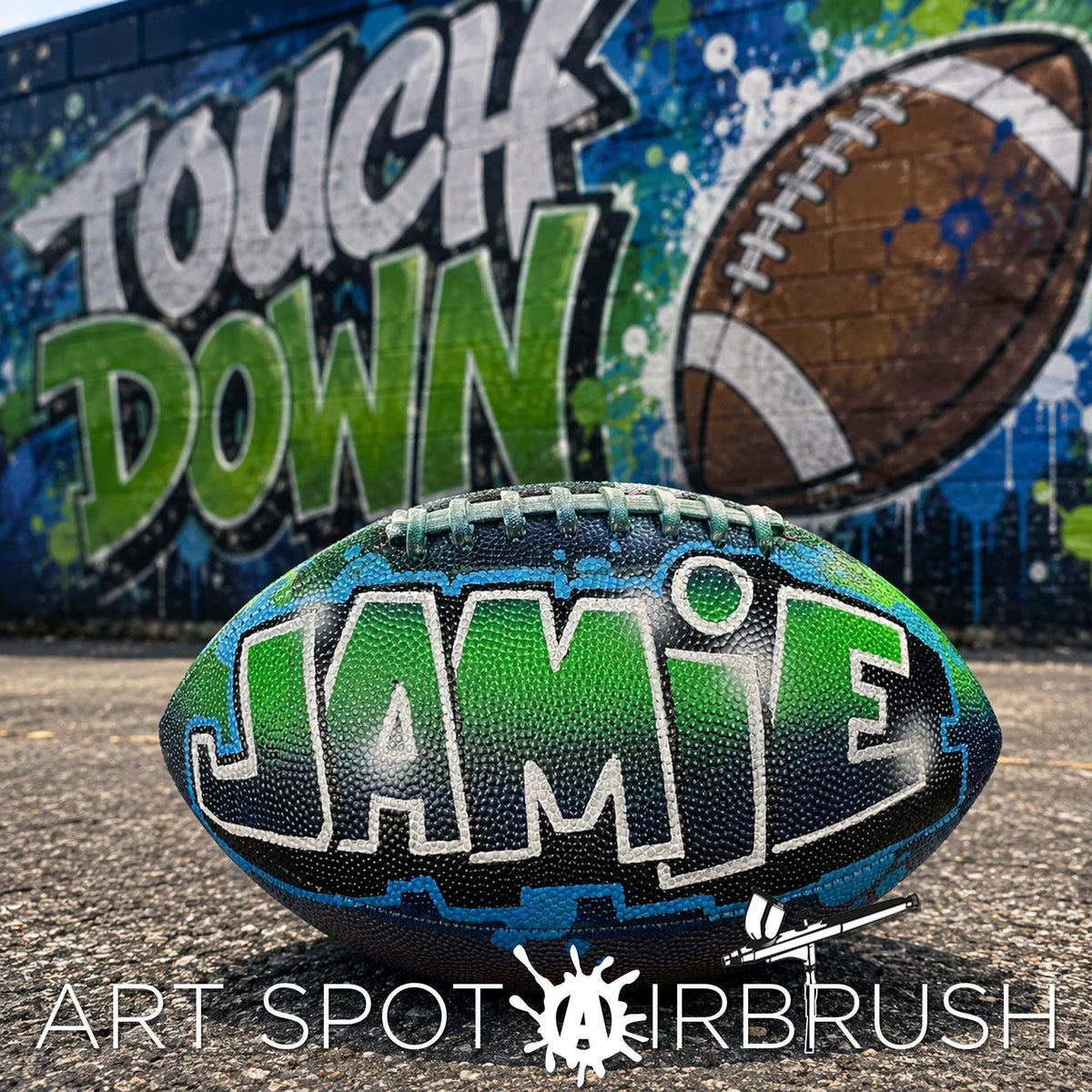A custom football personalized with the name "JAMIE" painted on it, placed in front of a graffiti-covered wall with the words "TOUCH DOWN" written on it.