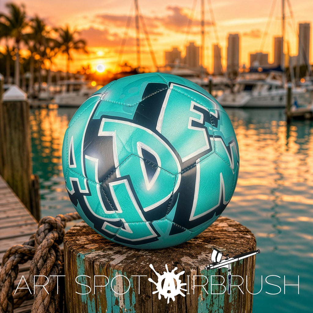 A teal-colored soccer ball with the word "ART" on it, placed on a wooden post near a body of water with boats and a sunset in the background.