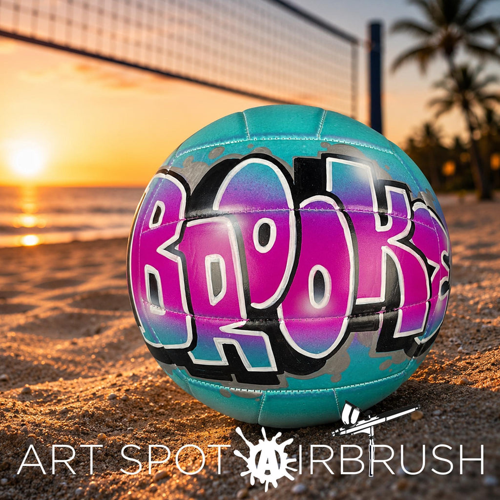 A colorful custom volleyball personalized with the word "Brooke" painted on it, resting on a sandy beach at sunset.