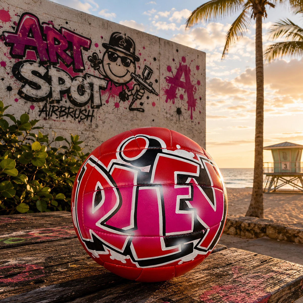 A red and black personalized custom volleyball with the word "RILEY" painted on it, sitting on a wooden surface near a graffiti-covered wall and palm trees.