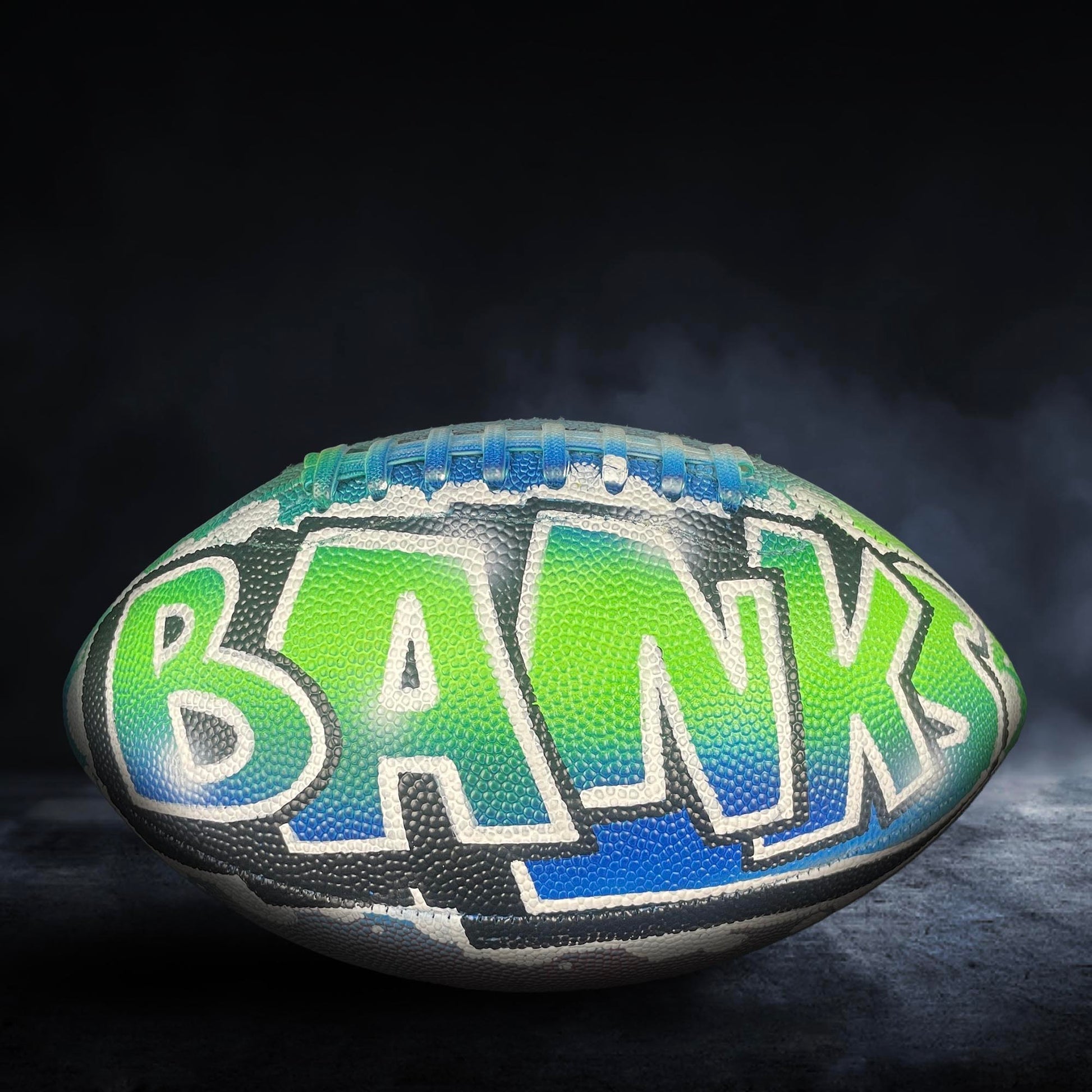 A football with the word "BANKS" painted on it, sitting on a dark surface with smoke or fog in the background.