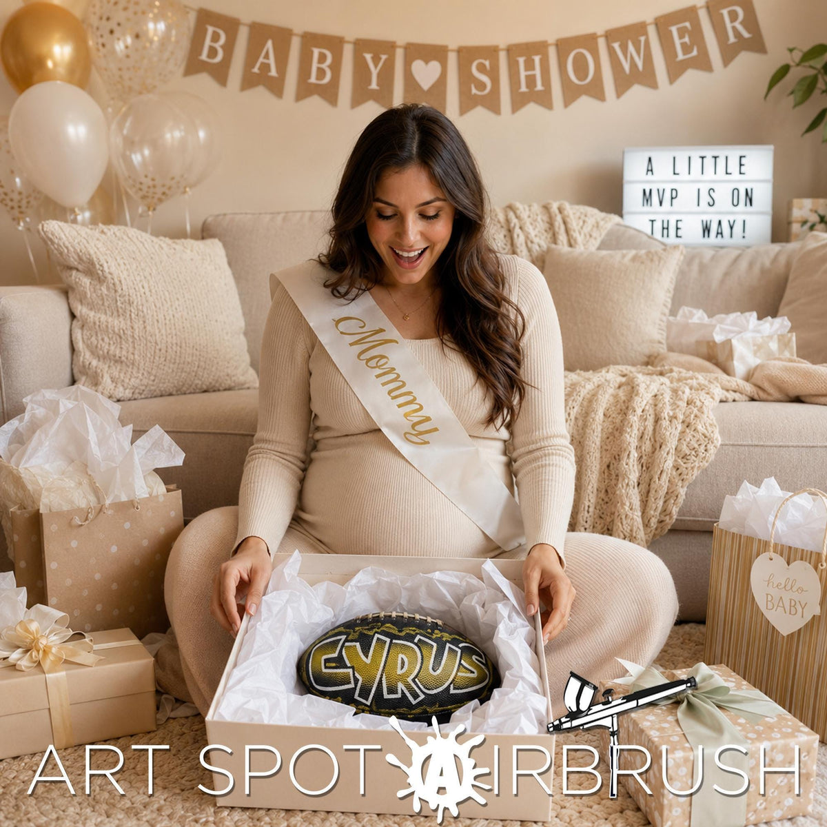 A pregnant woman is sitting on the floor surrounded by gifts, including a large box with apersonalized custom football with the name "CYRUS" and a sign that says "ART SPOT AIRBRUSH".