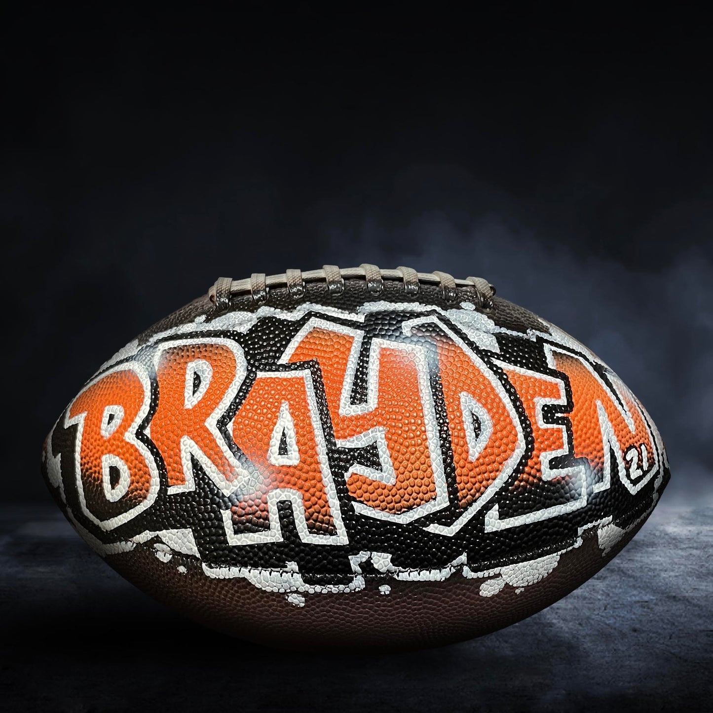 A football with the name "Brayden" painted on it, against a dark background with smoke.