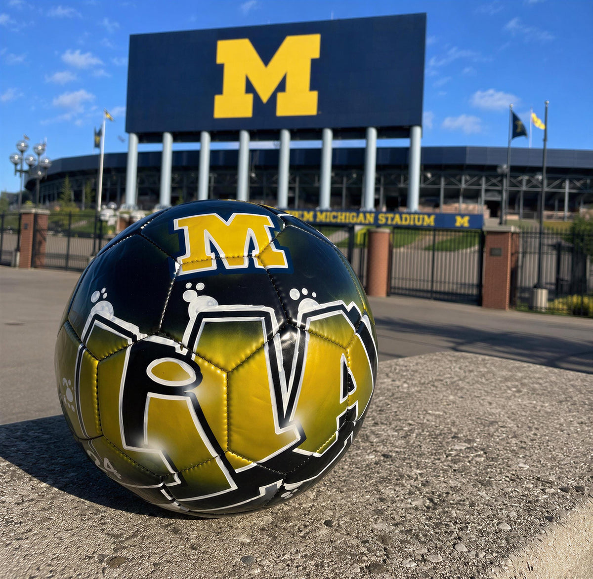 A soccer ball with the word "M" on it, placed on a concrete surface in front of a large stadium with a large "M" sign.