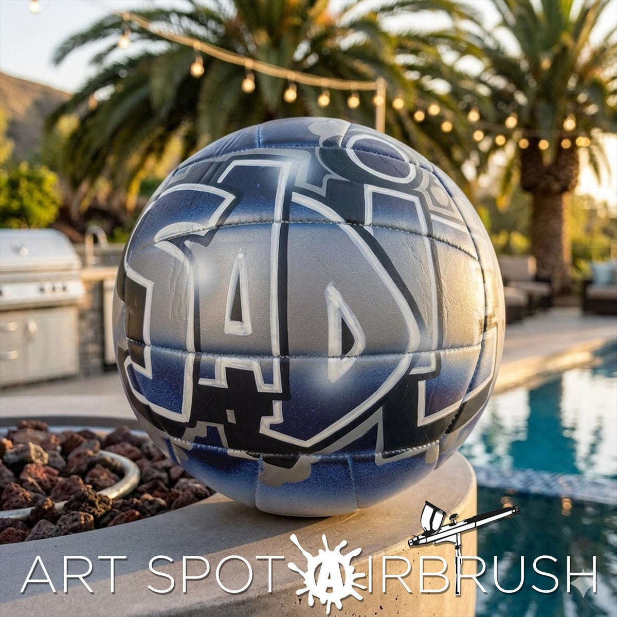 A blue and silver custom volleyball personalized with the word "SADIE" painted on it, sitting on a ledge near a pool and palm trees.
