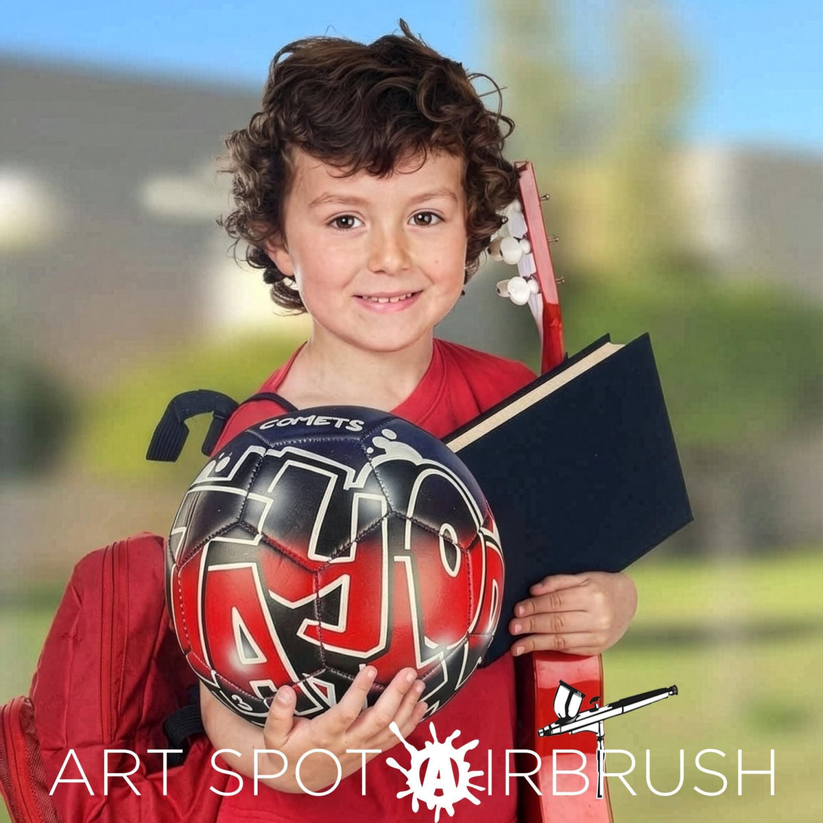 A young boy with curly hair is smiling and holding a red and black soccer ball, a black book, and a red guitar.