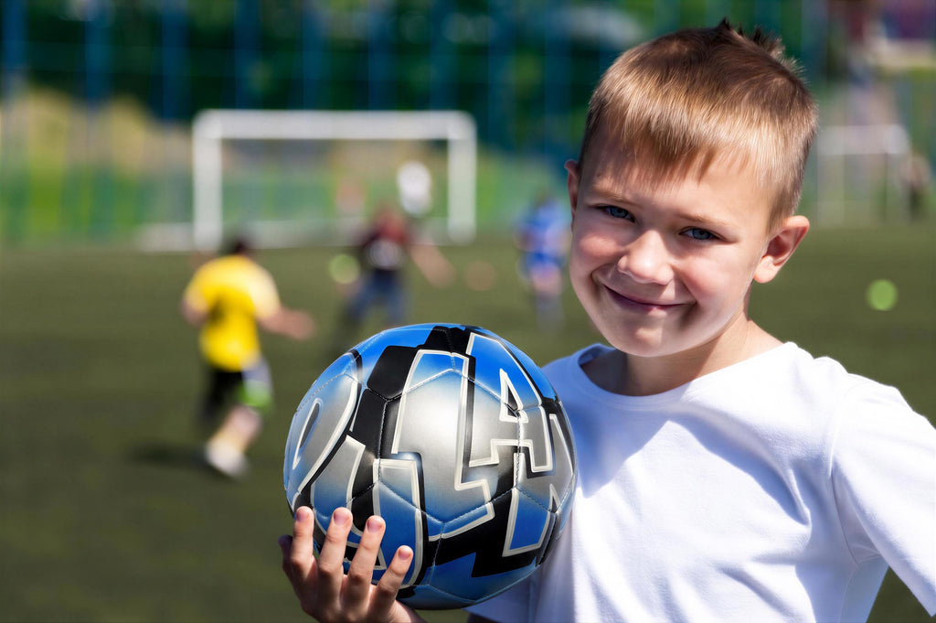 A young boy is holding a soccer ball and smiling at the camera, with other children playing soccer in the background.