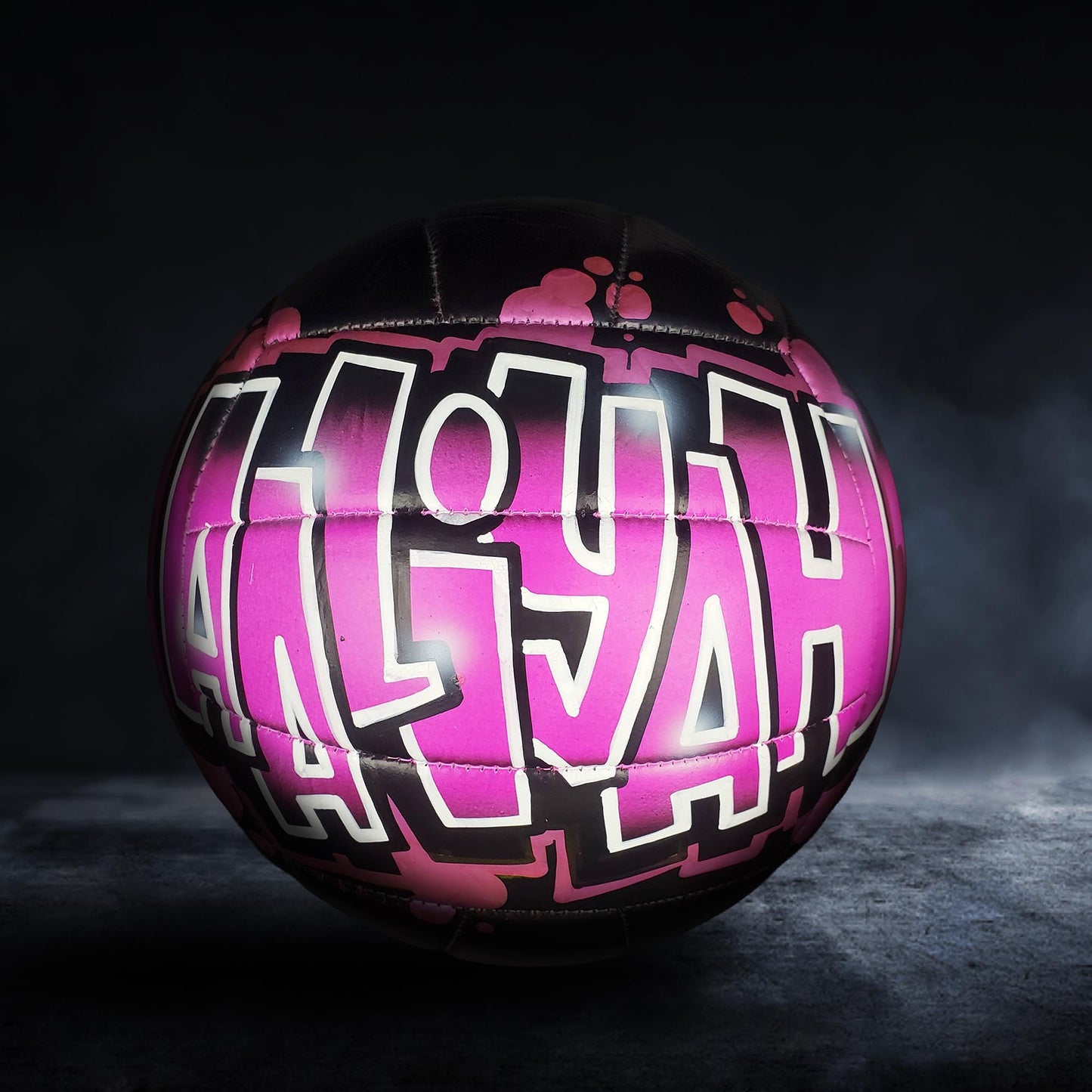 A pink and black soccer ball with the name "ALIH" painted on it, against a dark background.