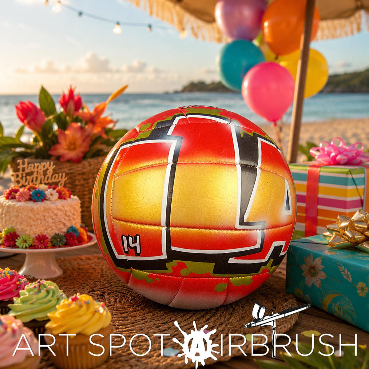 A birthday celebration with a custom volleyball personalized with a name, cake, gifts, and balloons set up on a beach, with a clear blue sky and ocean in the background.