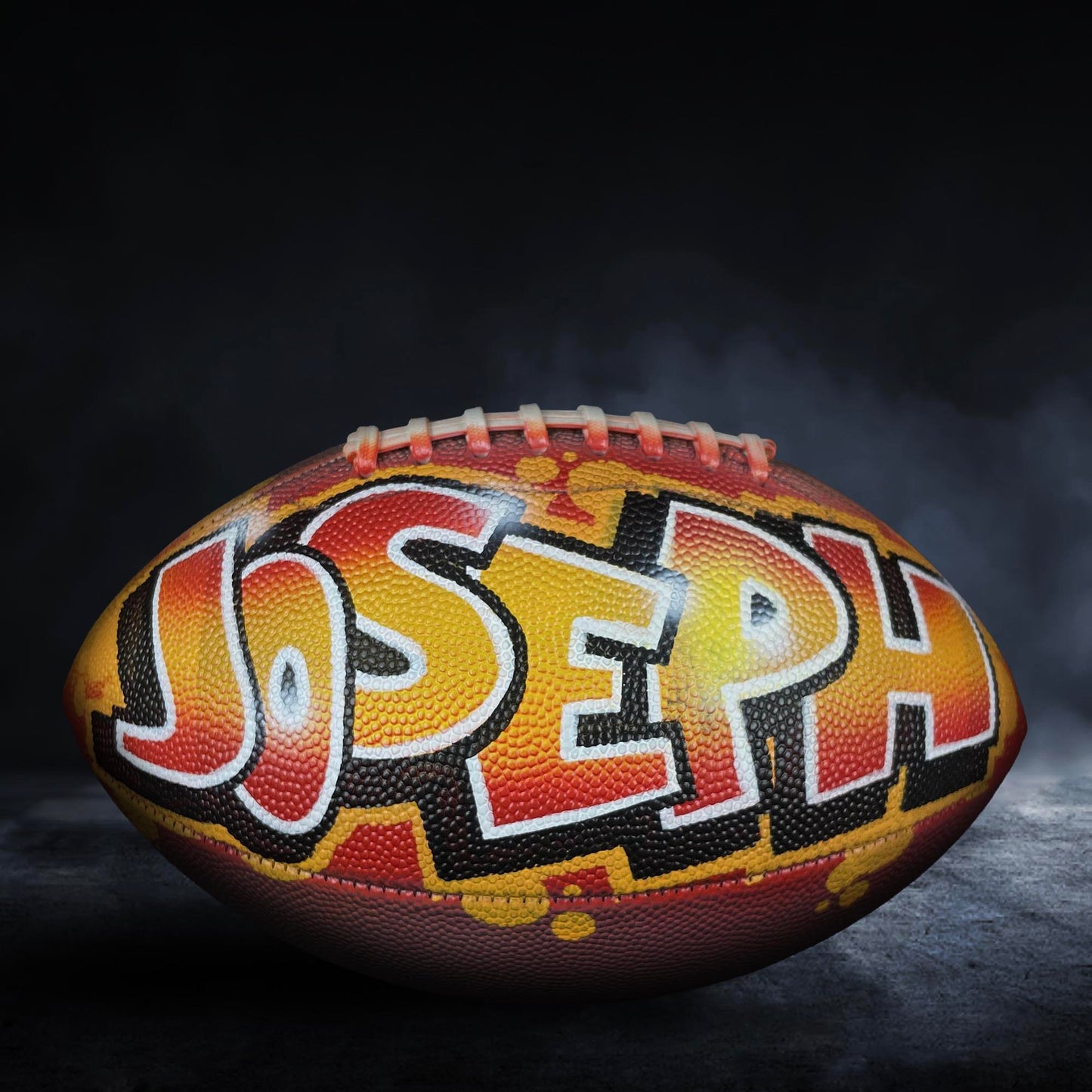 A football with the name "Joseph" painted on it, set against a dark background with smoke or fog.