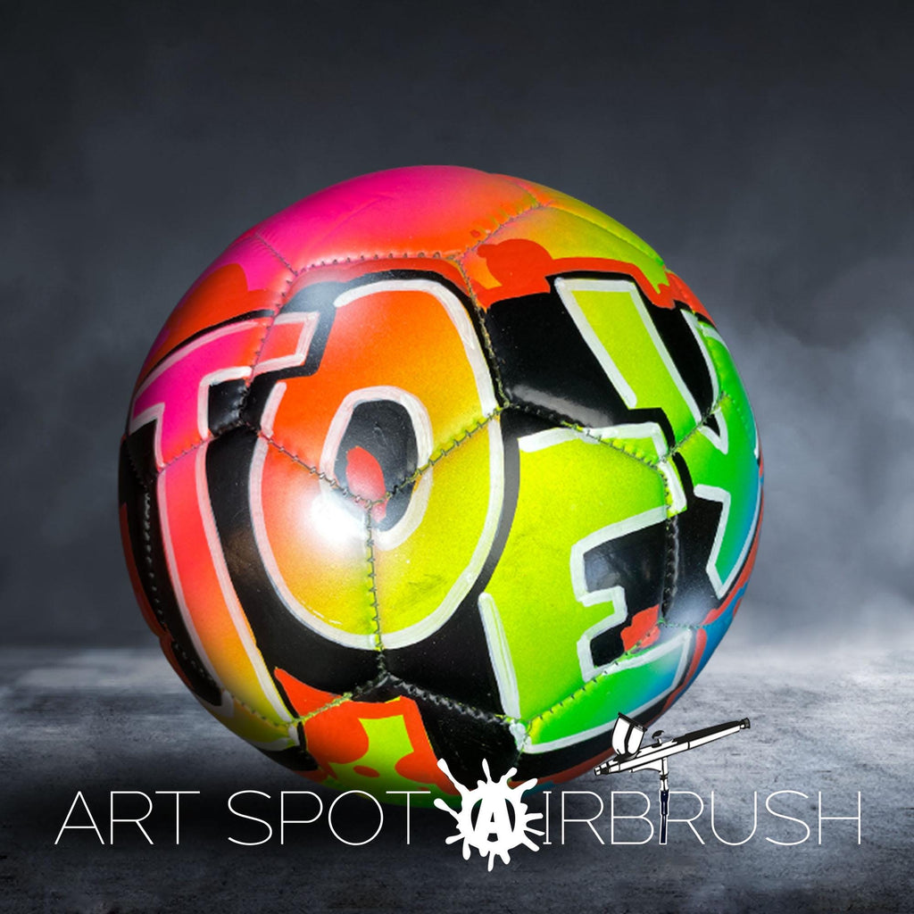 A colorful, graffiti-style soccer ball with the word "TOY" painted on it, set against a dark background.