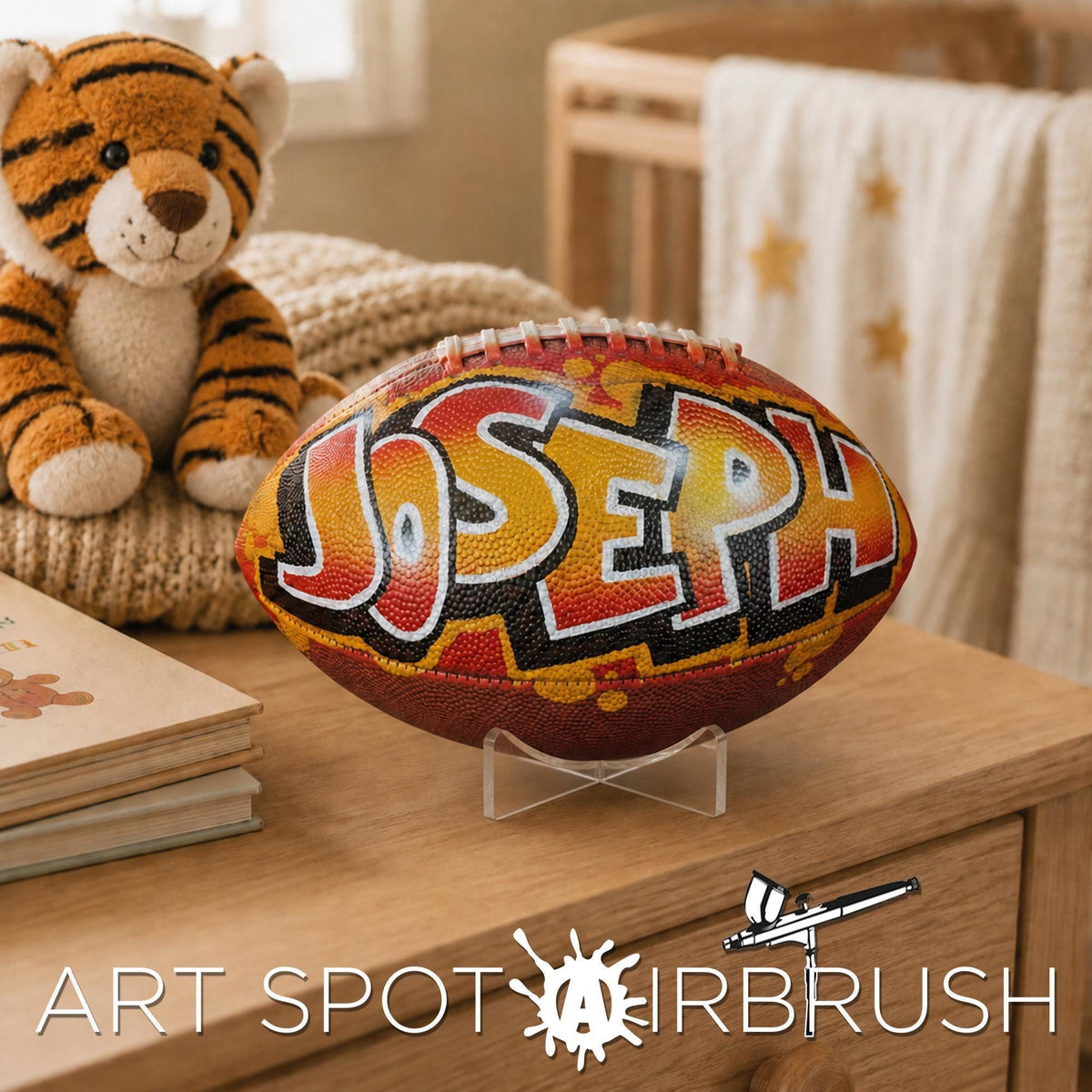 A personalized football with the name "Joseph" painted on it, sitting on a wooden surface next to a stuffed tiger toy and some books.
