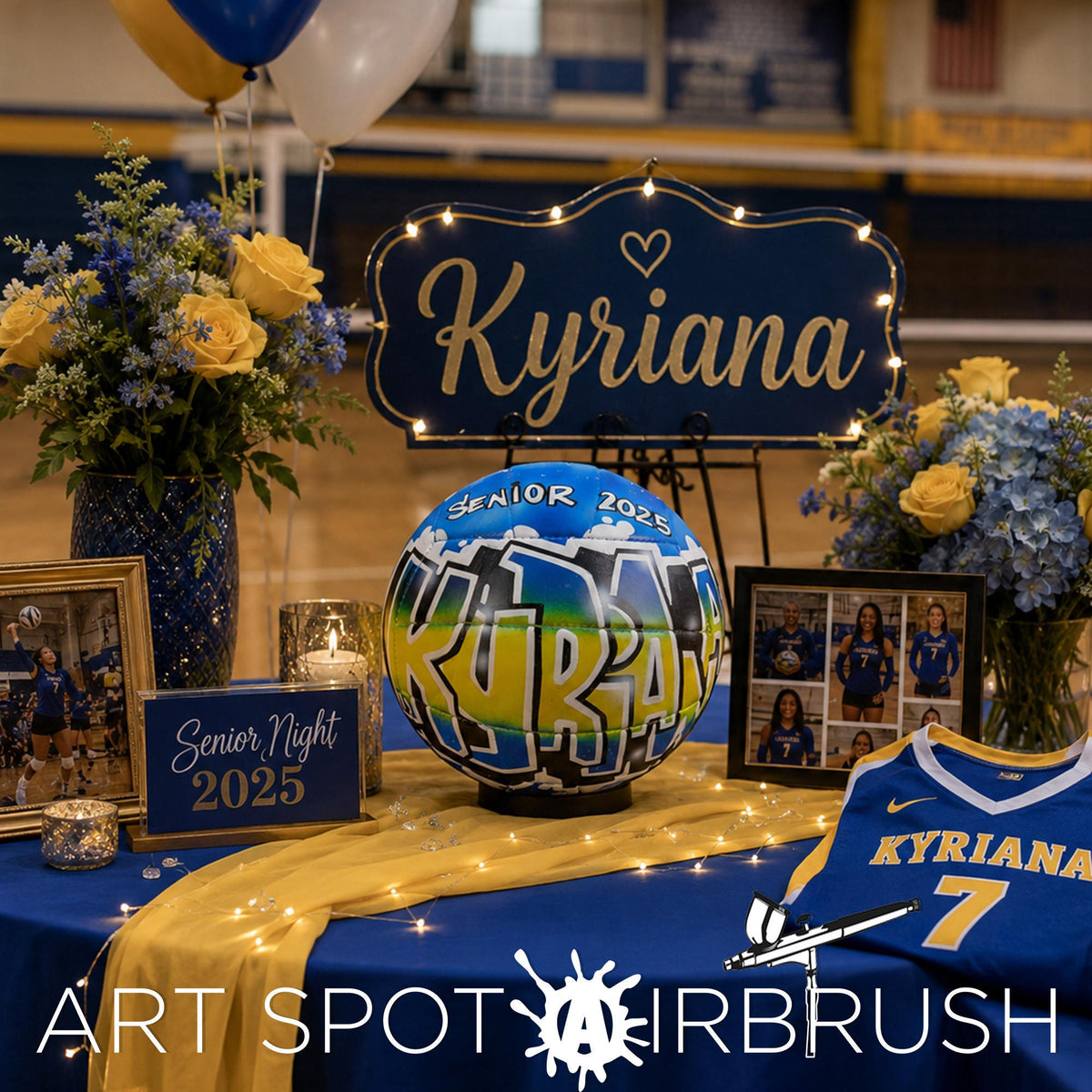 a graduation celebration with a blue and gold color scheme. The centerpiece is a large sign that says "Kyriana" in gold lettering. Surrounding the sign are various decorations, including balloons, flowers, and framed photos. A custom volleyball personalized with the word "Kyriana" on it is also present. The table is covered with a blue tablecloth and has a sign that reads "Art Spot AIRBRUSH".