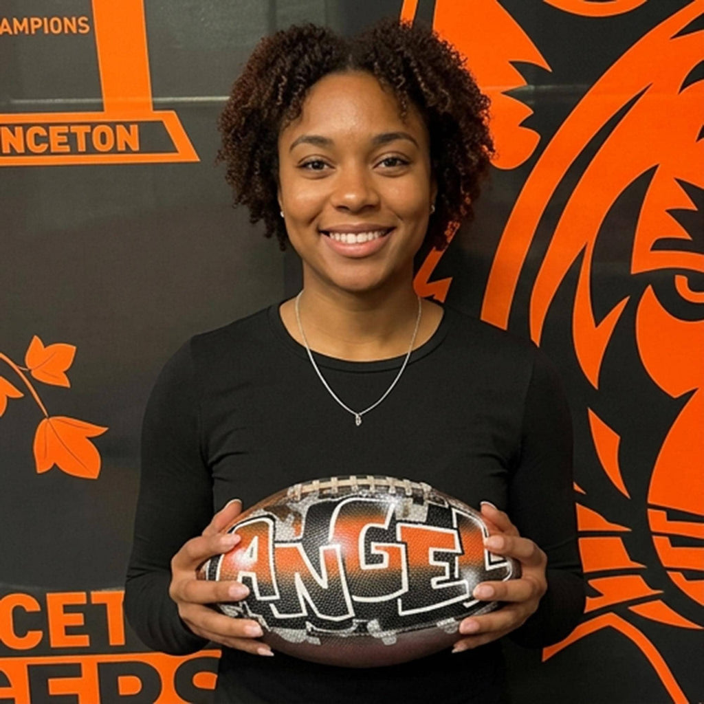 A woman is holding a custom football personalized with the word "ANGEL" written on it, smiling in front of a large orange and black banner.