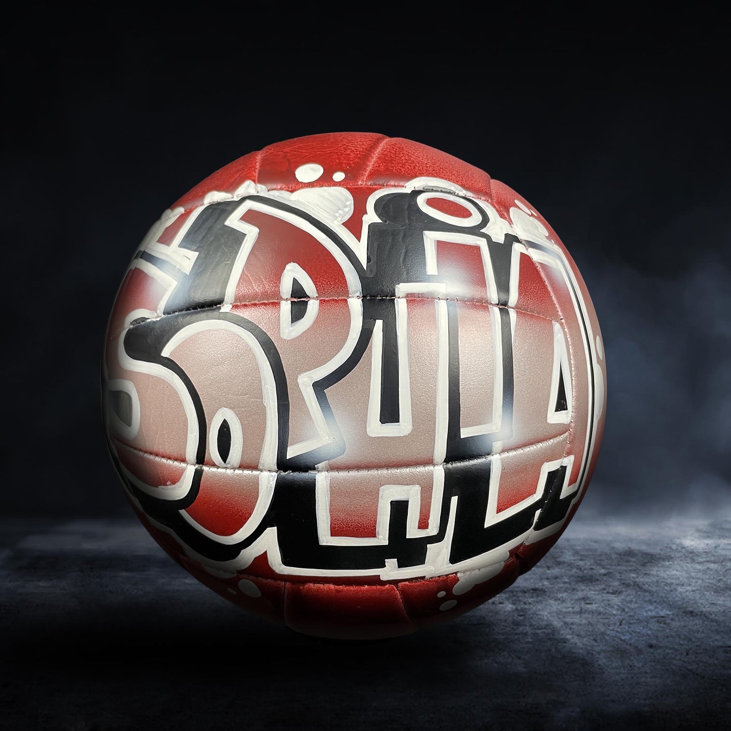 A red and black soccer ball with graffiti-style text on it, set against a dark background.