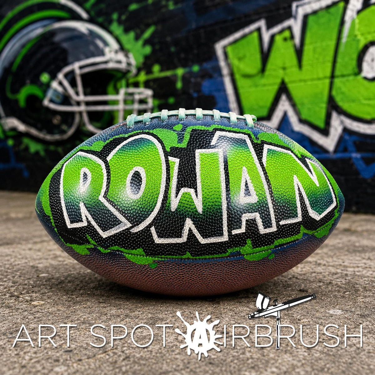 A green and black custom football personalized with the name "Rowan" painted on it, placed on a concrete surface in front of a graffiti-covered wall.