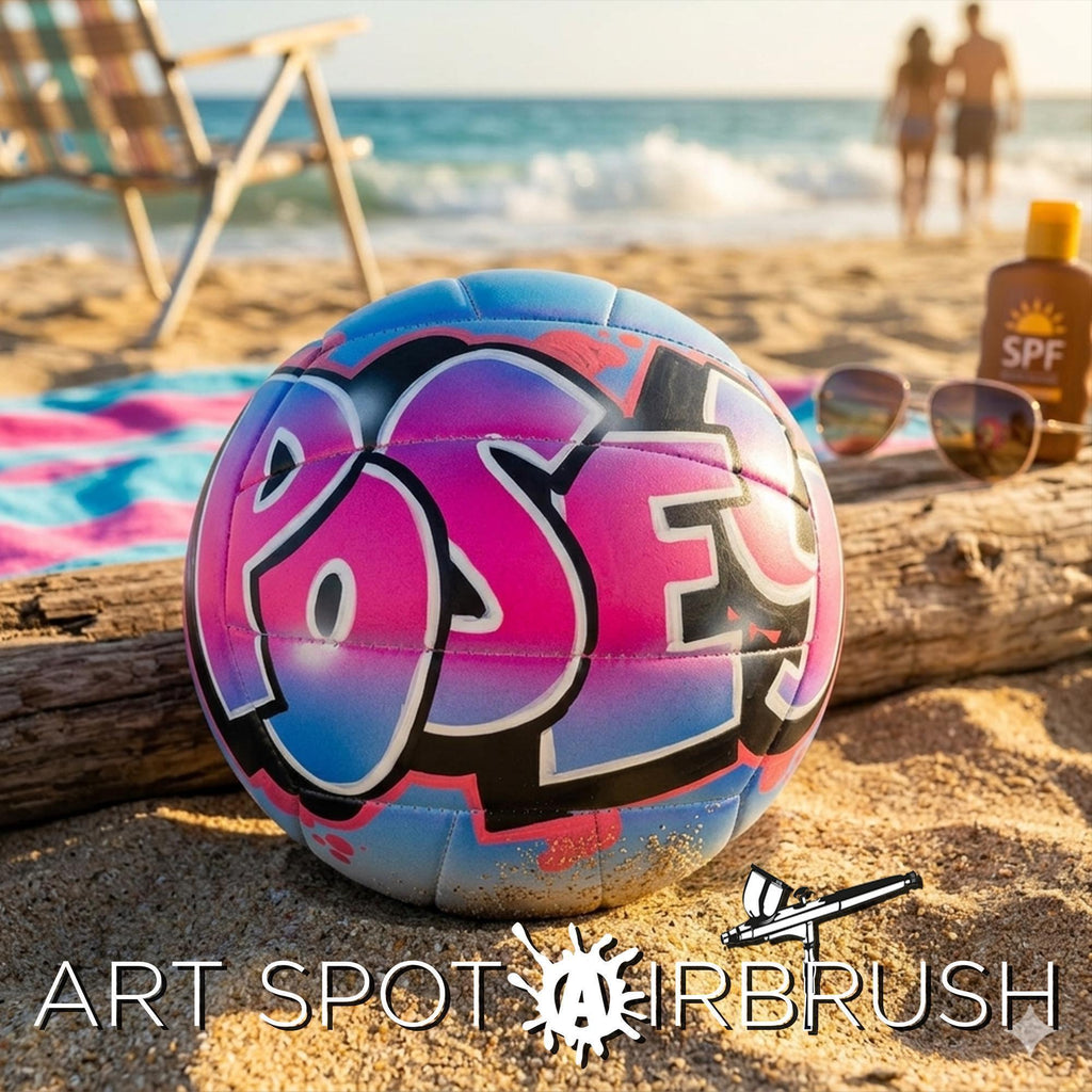 A custom volleyball with graffiti-style writing on it, placed on a sandy beach with a chair and sunglasses in the background.