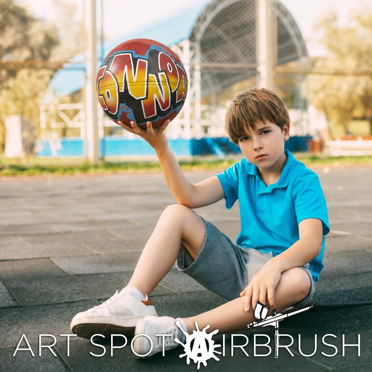 A young boy in a blue shirt sits on the ground, holding a colorful sports ball with graffiti on it.