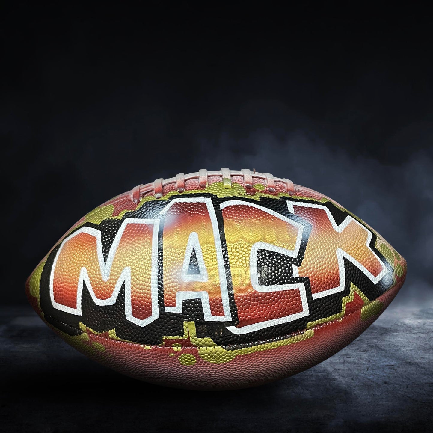 A football with the name "MACK" painted on it, sitting on a dark surface with smoke or fog in the background.