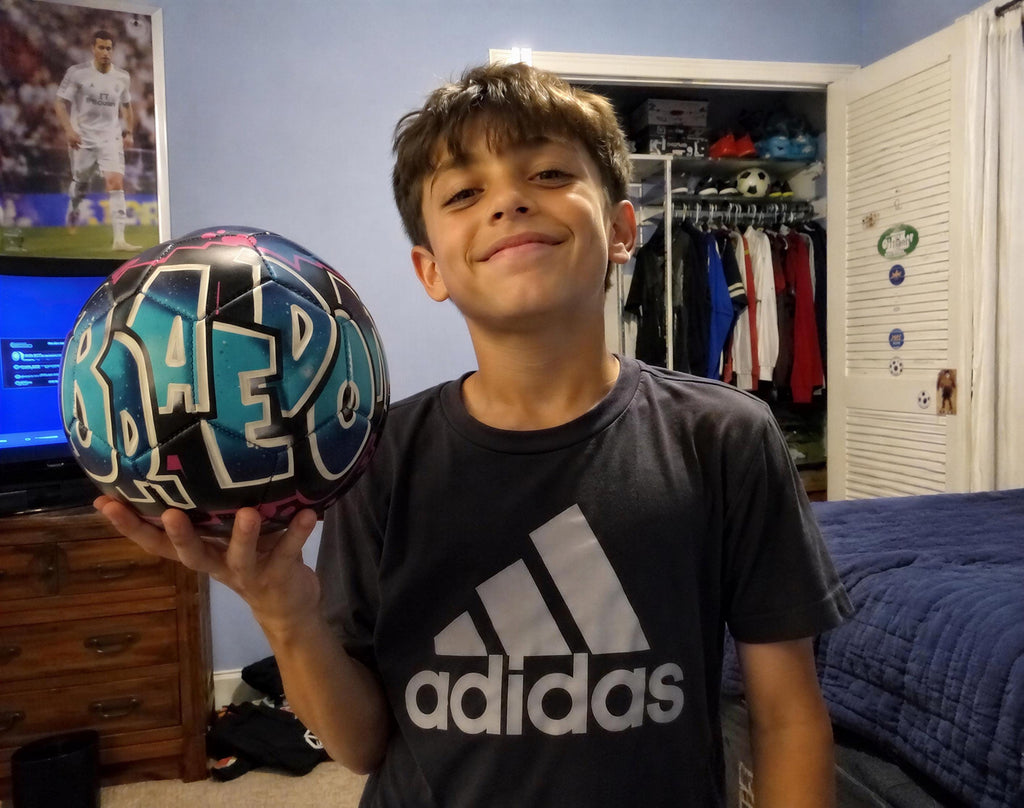 A young boy is holding a soccer ball with the word "BARO" on it, smiling and posing in a bedroom.