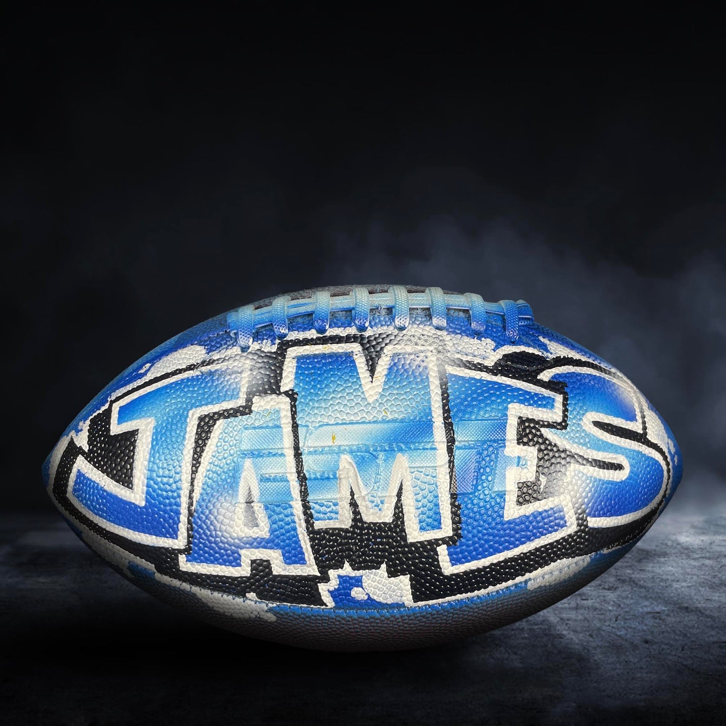 A blue and black football with the name "JAMES" painted on it, sitting on a dark surface with smoke or fog in the background.