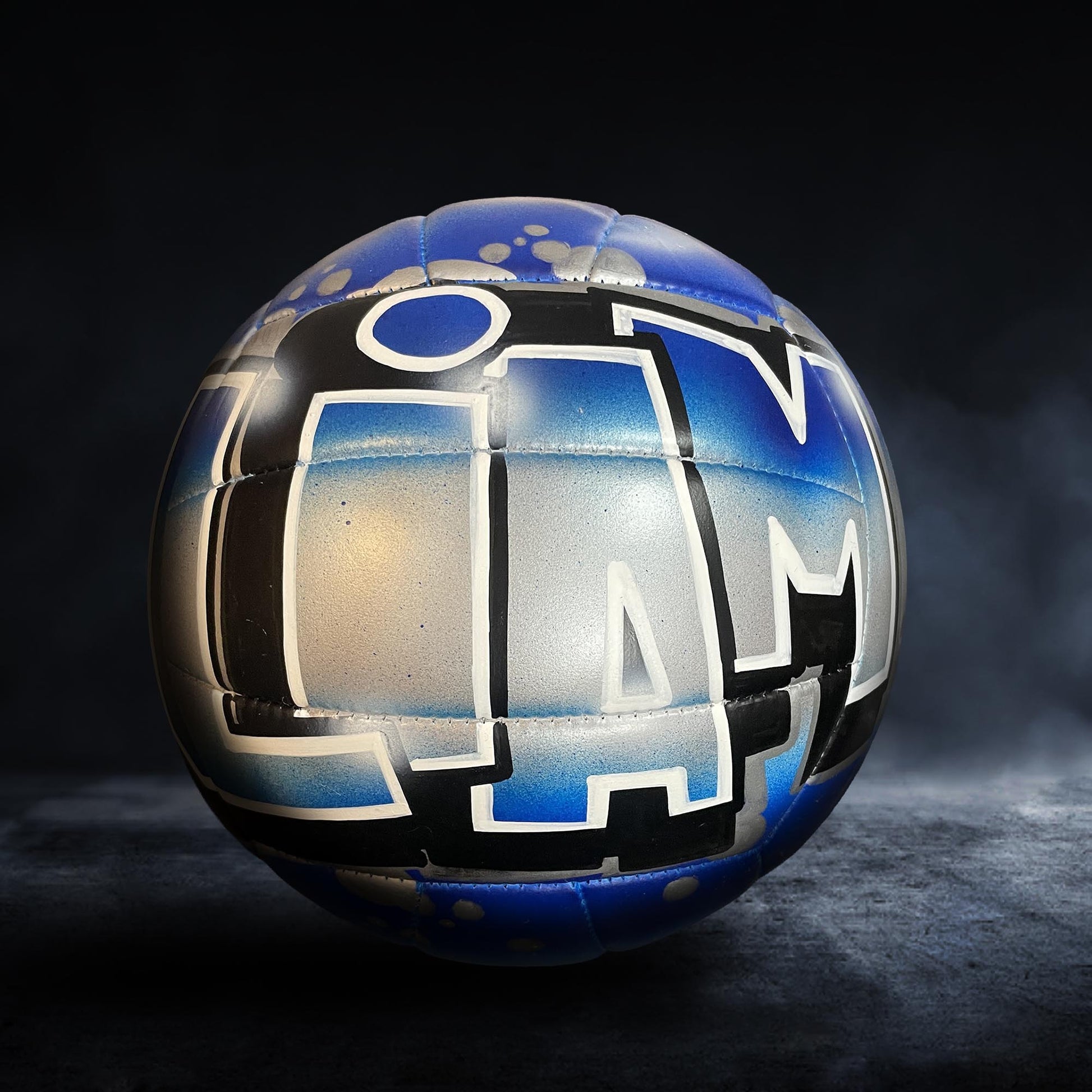 A blue and silver soccer ball with the word "LAM" painted on it, sitting on a dark surface.