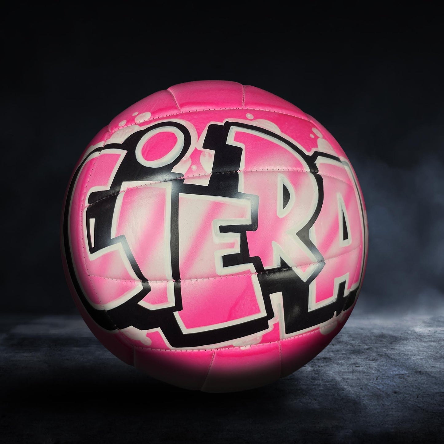 A pink and black volleyball with graffiti-style text on it, sitting on a dark surface.
